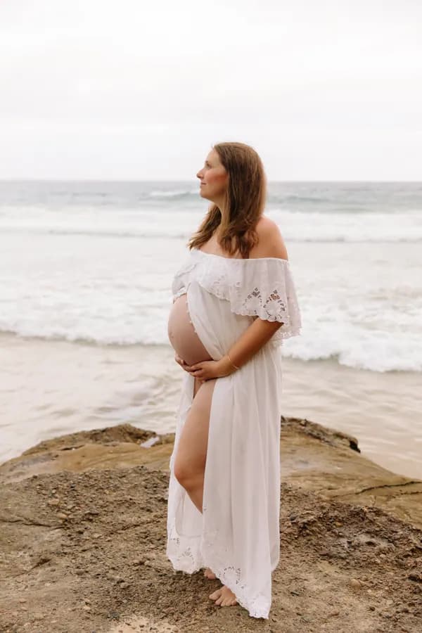 Courtney in her fourth pregnancy, standing on rocks by the sea in a white dress